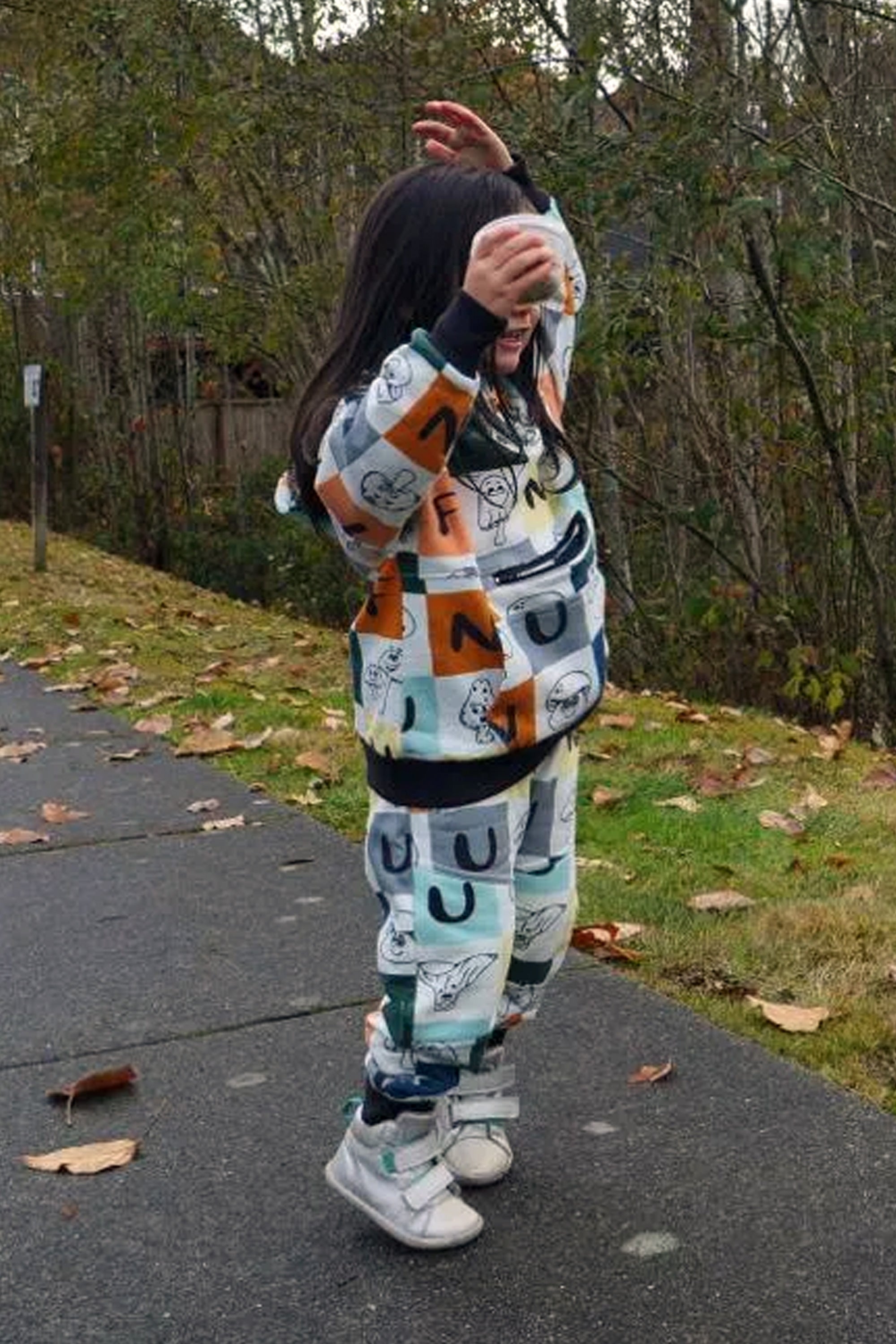 A child wearing a matching two-piece Hoodie and Joggers with a colorful Fungi print mushroom andi letters, standing on an outdoor paved path.