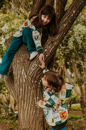 Two children wearing matching mushrooms fungi-patterned hoodie and teal pants play on a large tree trunk in a park setting.