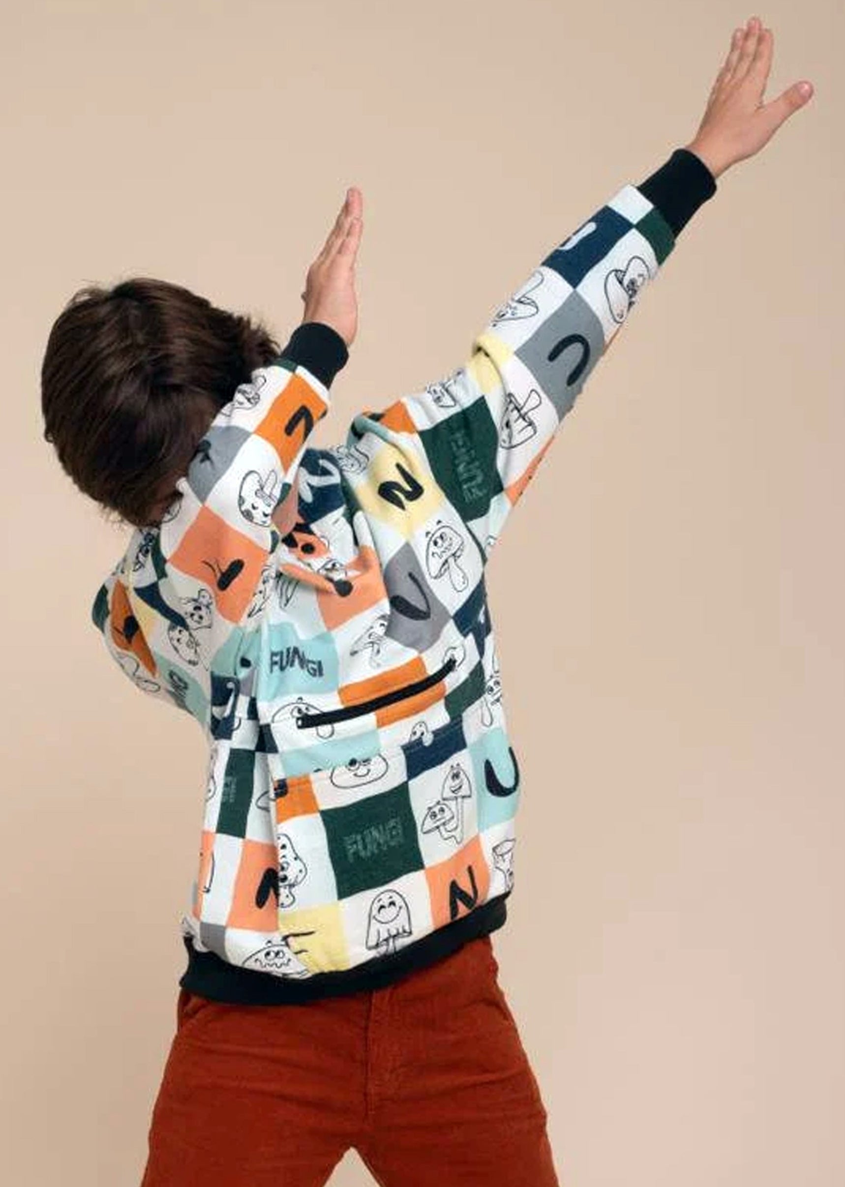 A child model from behind wearing a colorful fungi mushrooms patterned hoodie, rust-brown corduroy pants, posing with arms raised against a solid beige studio background.