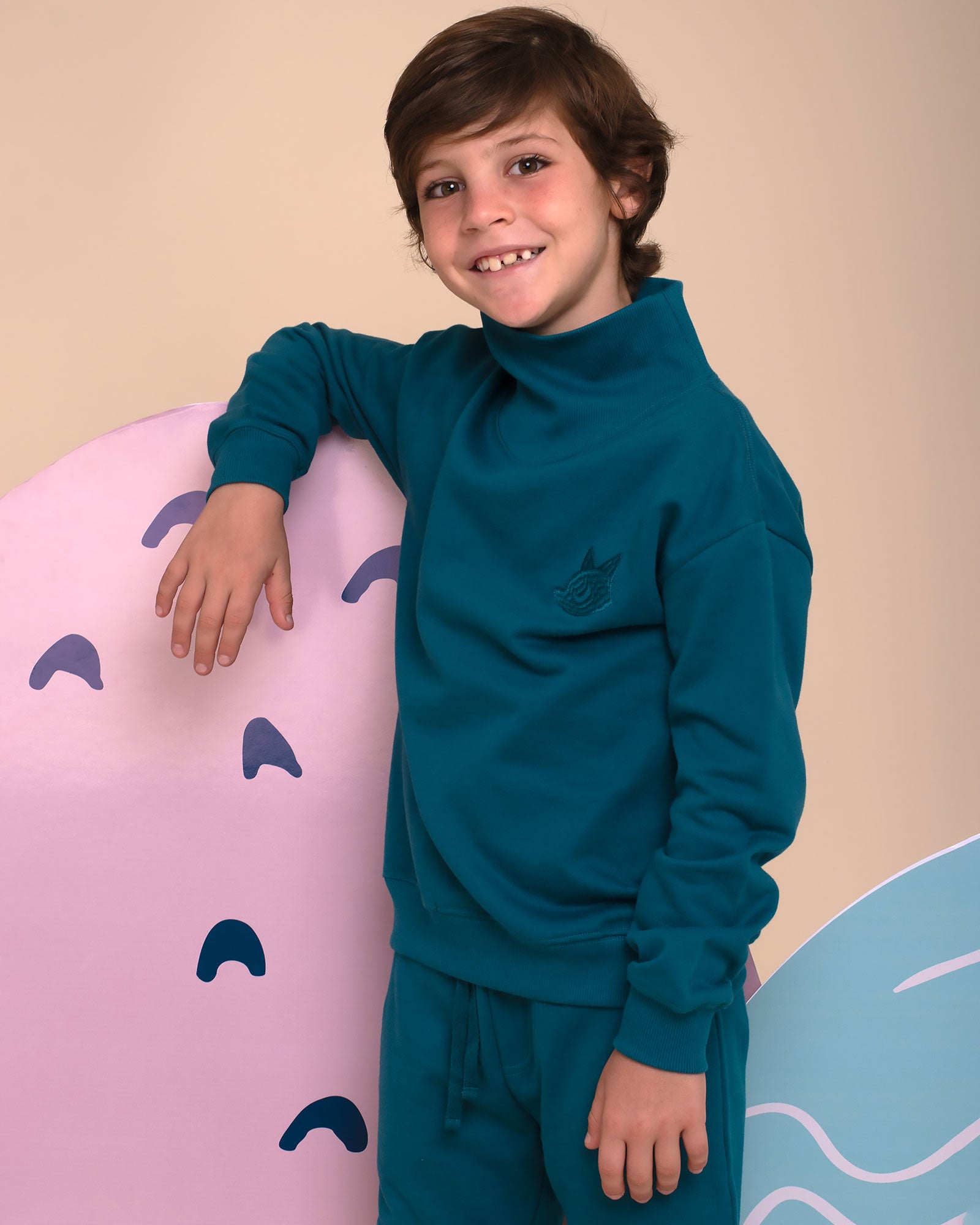 Child wearing a blue funnel neck sweatshirt and matching joggers outfit standing against a beige background.