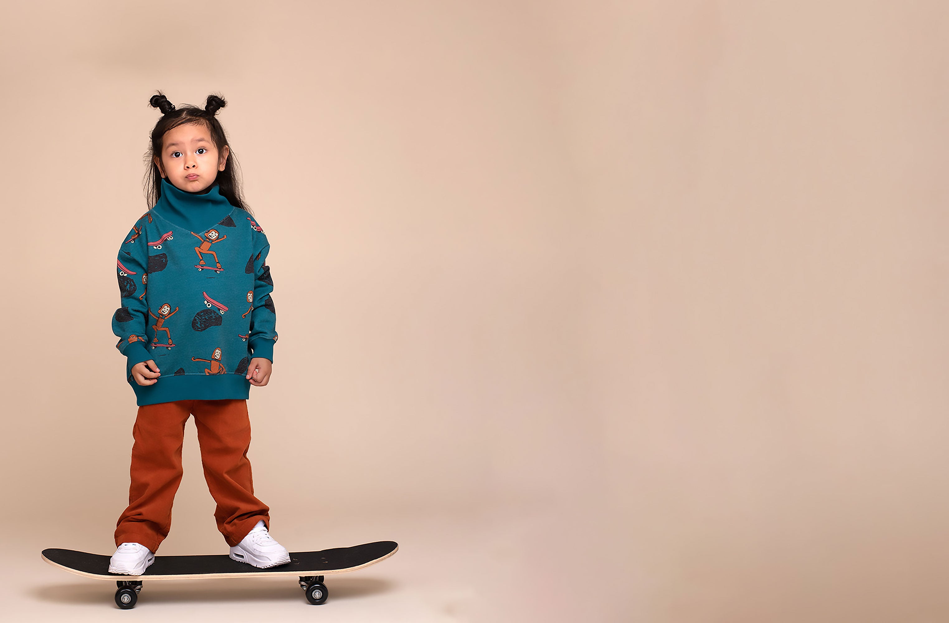 Child standing on a skateboard in a studio, wearing a blue Rapozza skater monkey print sweatshirt with brown pants against a neutral background.
