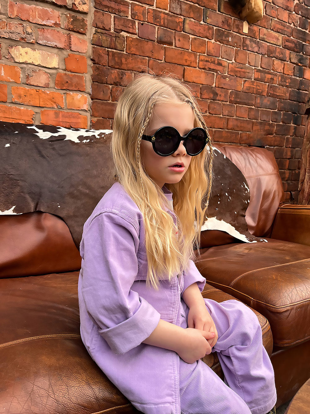 Young girl with long blonde hair wearing oversized round sunglasses and a lilac corduroy jumpsuit, sitting on a leather sofa in front of a brick wall.