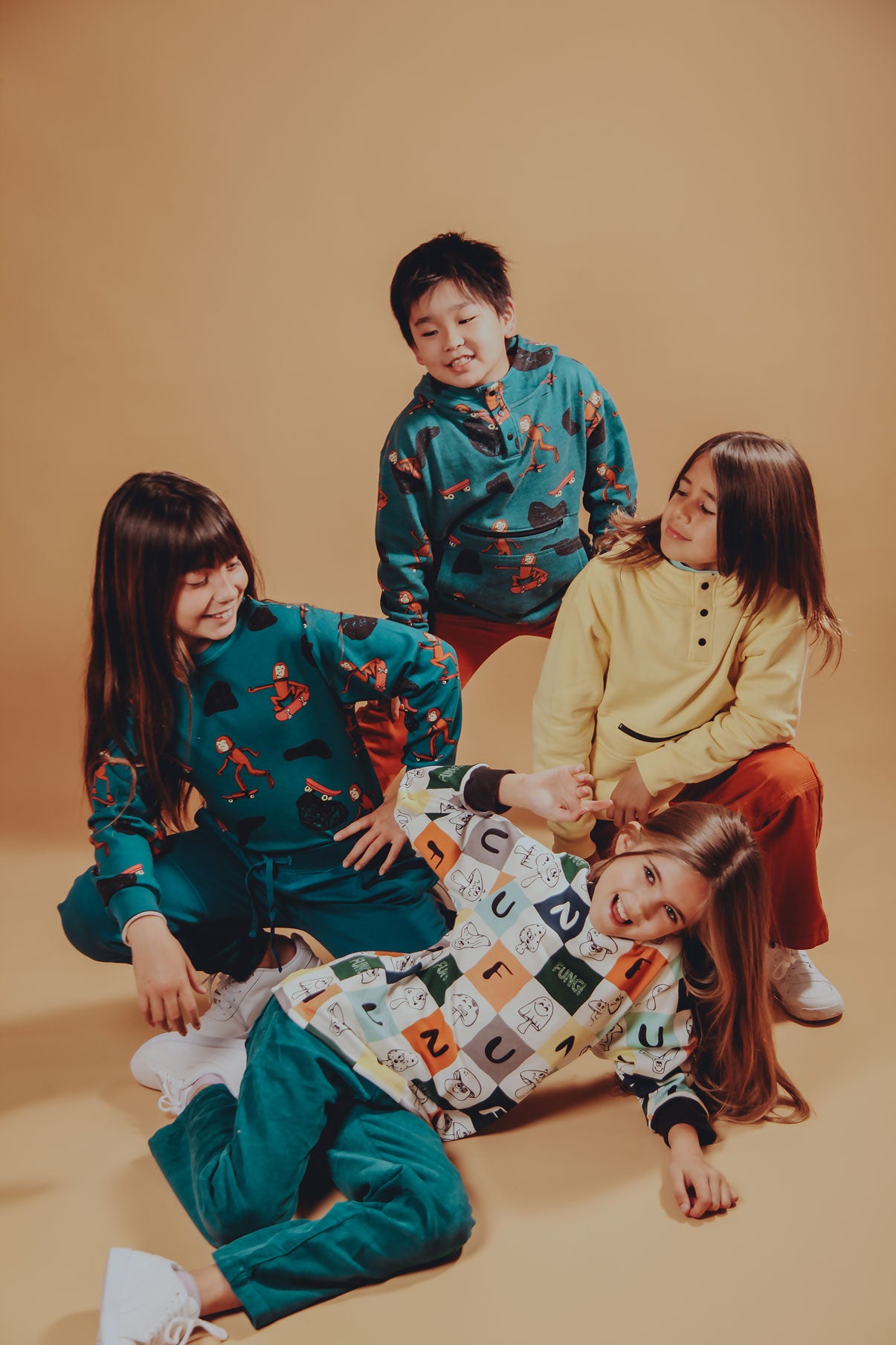 Four children laughing and playing in a warm studio, wearing colorful Rapozza sweatshirts including fungi and skater monkey prints with cozy corduroy pants