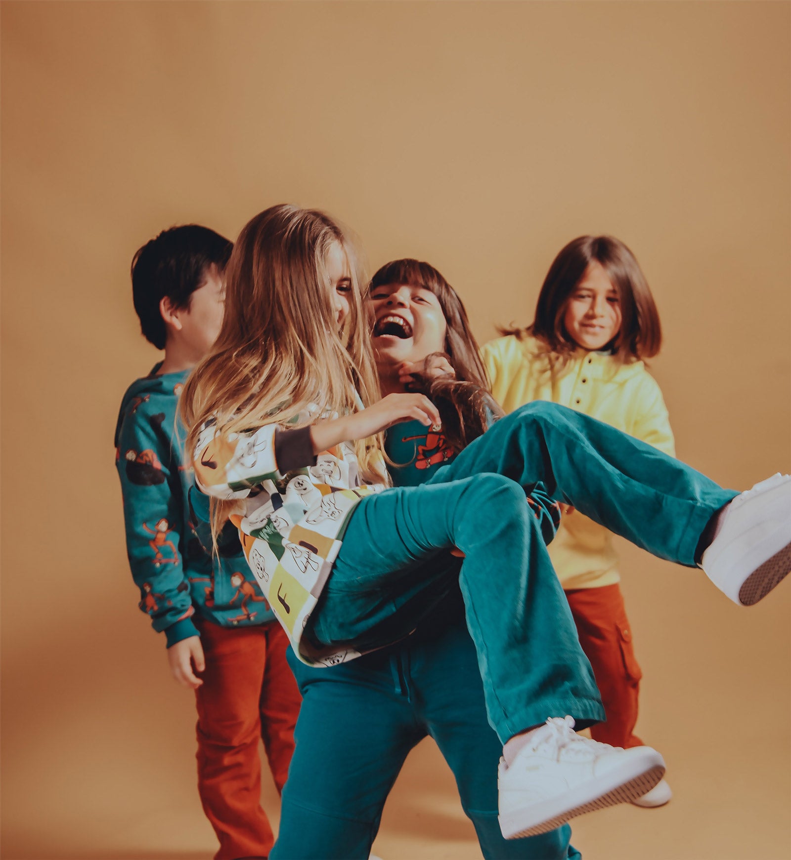 Group of children laughing and playing in a warm studio, one girl lifting another while they all wear colorful Rapozza sweatshirts and joggers