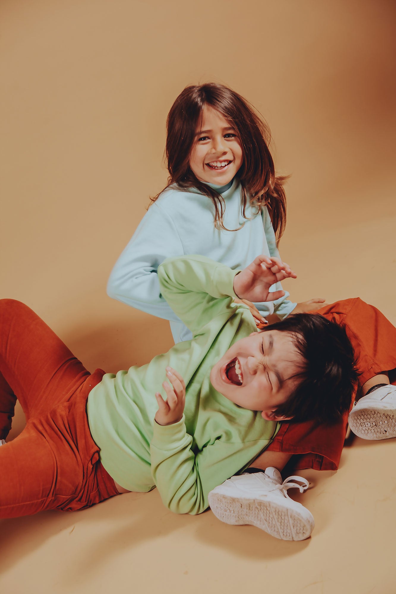 Two children laughing and playfully tumbling on the studio floor, wearing Rapozza colorful long-sleeve basics with brown corduroy pants